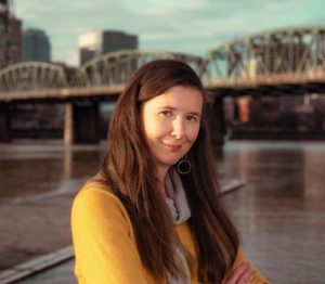 Haven Wheelock, a woman with long brown hair and a yellow sweater, stands in front of the Willamette River with a bridge in the background