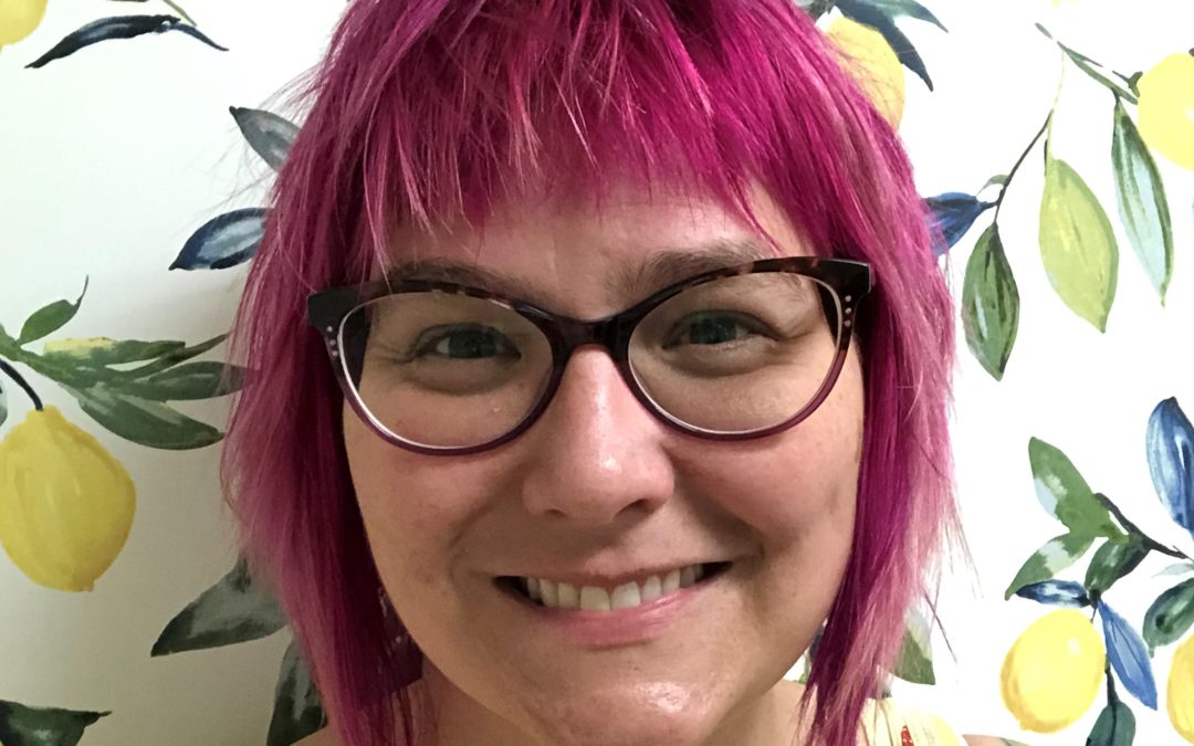 Housing Program Manager, Lindsay, has pink hair, glasses, and is smiling in front of wallpaper with brightly colored fruit trees