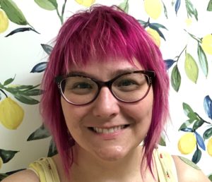 Housing Program Manager, Lindsay, has pink hair, glasses, and is smiling in front of wallpaper with brightly colored fruit trees