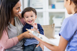 A woman wearing a striped shirt and sweater is holding a young child who is smiling. They are talking to a medical provider who has their back to the camera