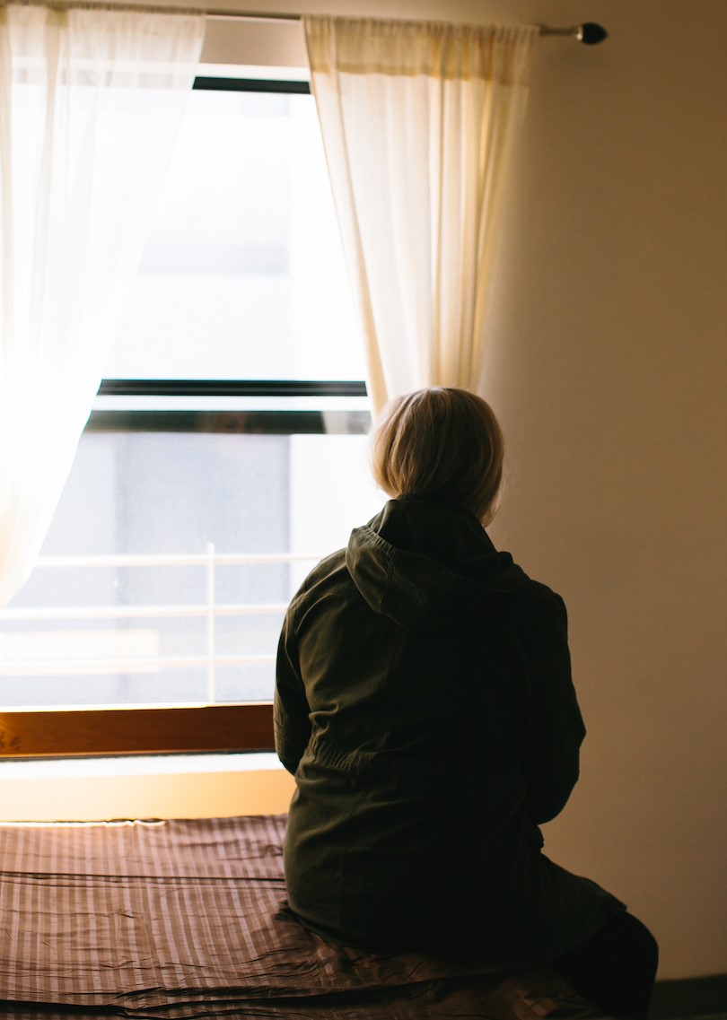 Young person sitting on a bed looking out the window, their face cannot be seen