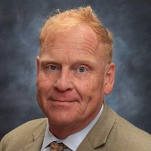 Don Reuther, a human with light hair wearing a tan suit and tie. The image is a headshot.