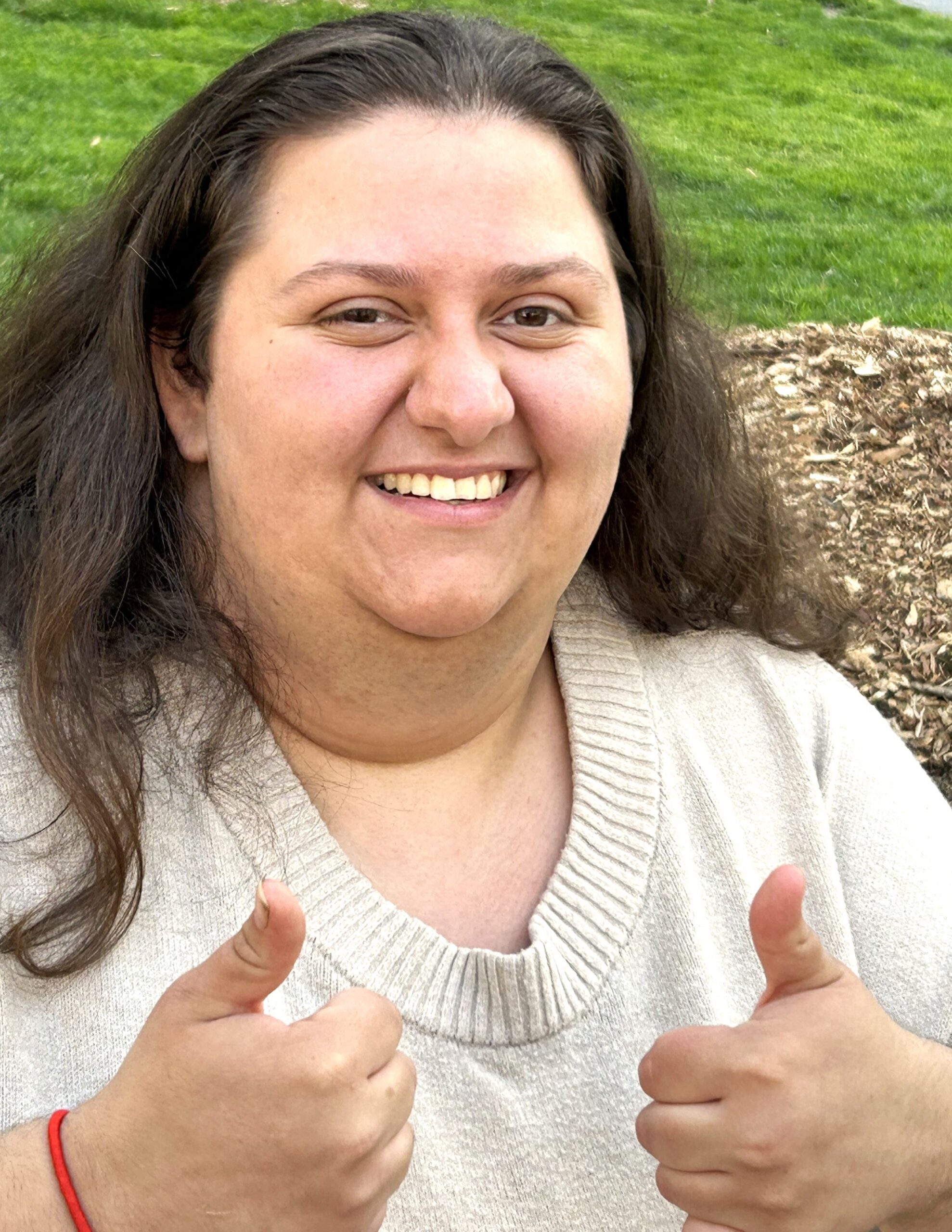 Tea, age 23, an Outside In participant with long brown hair stops at a green park, smiling broadly and gestures with two thumbs up.